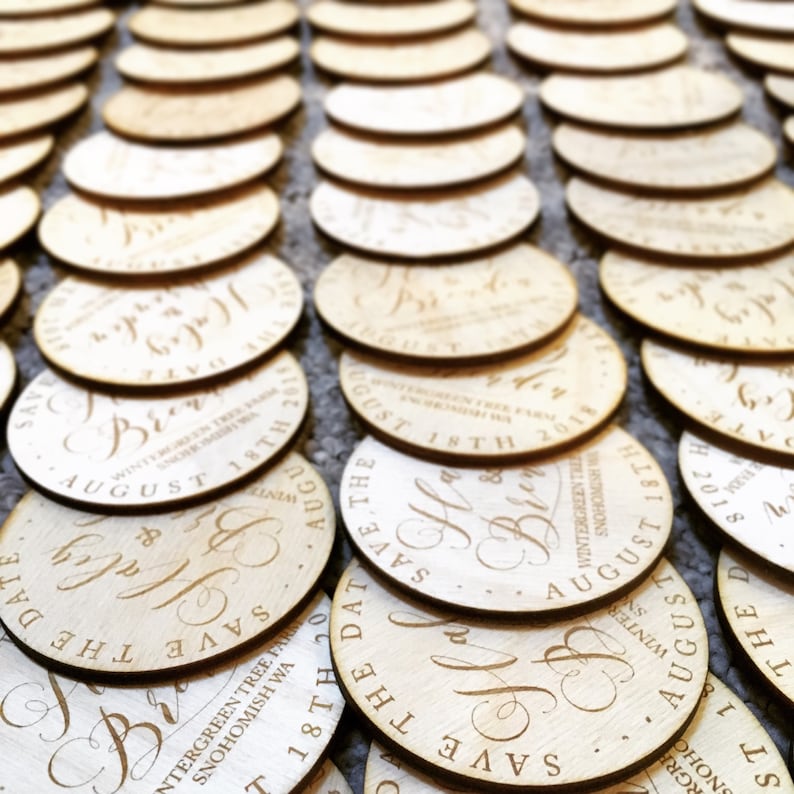 May include: A collection of round, wooden save the date cards with calligraphy writing. The cards are engraved with the text "Save the Date" and the date "August 18th, 2019".