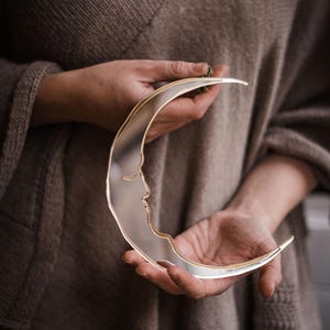 May include: A silver crescent moon shaped mirror with a face design in the centre. The mirror is being held by a person wearing a brown sweater.