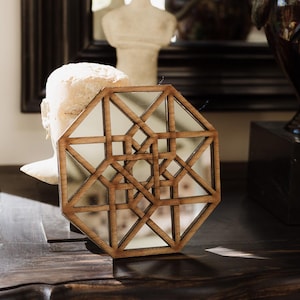 May include: Geometric wooden mirror with a complex, symmetrical design. The mirror is octagonal with a series of intersecting lines creating a pattern. The frame is made of light brown wood. The mirror is placed on a dark wooden surface.