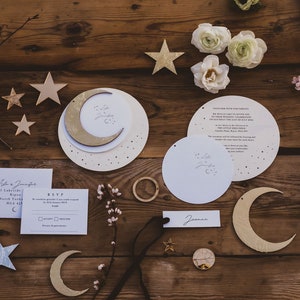 Könnte beinhalten: Eine Hochzeitseinladungssuite mit einem himmlischen Thema. Die Einladung zeigt einen goldenen Halbmond und Sterne auf weißem Hintergrund. Die RSVP-Karte ist weiß mit goldfarbenem Rand. Die Einladung befindet sich auf einem hölzernen Hintergrund mit weiteren himmlischen Dekorationen.