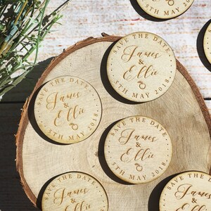 May include: Five round wooden save the date magnets with the text "Save the Date" and the names "James & Ellie" and the date "5th May 2018".