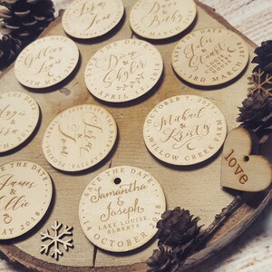 May include: A collection of round wooden save the date cards with calligraphy lettering. The cards are decorated with a variety of designs, including hearts, snowflakes, and pine cones. The text on the cards includes the names of the couples, the date of the wedding, and the location.