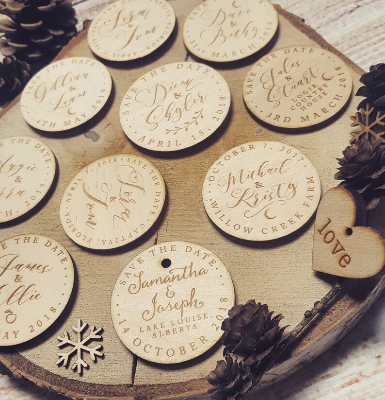 May include: A collection of round wooden save the date cards with calligraphy lettering. The cards are decorated with a variety of designs, including hearts, snowflakes, and pine cones. The text on the cards includes the names of the couples, the date of the wedding, and the location.
