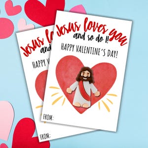 May include: Two Valentine's Day cards with the message "Jesus loves you and so do I! Happy Valentine's Day!" printed in red. Each card features a red heart and an illustration of Jesus. Pink and red paper hearts are scattered around the cards.
