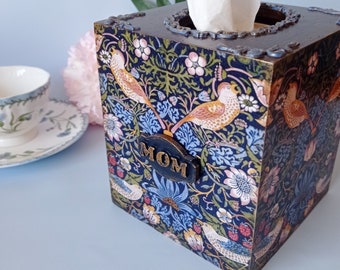 Puede incluir: Una tapa de caja de pañuelos de madera oscura con un patrón floral en tonos azules, verdes y rosas. La tapa tiene una pequeña placa de metal que dice "MOM".