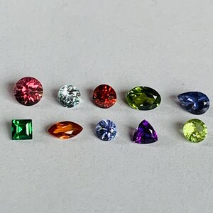 May include: An assortment of colorful gemstones in various shapes, including round, oval, and square. The gems display a range of colors, such as red, green, blue, and purple. The gemstones are arranged on a white surface.