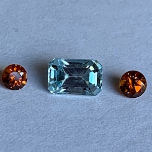 May include: Three gemstones: a light blue emerald cut gemstone, and two round orange gemstones.