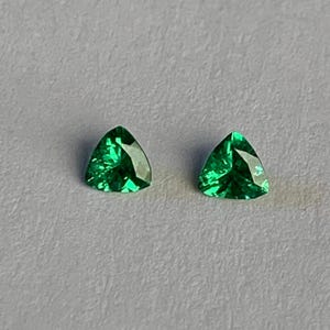 May include: Two loose, vibrant green gemstones cut into a triangular shape. Each stone features a faceted surface that reflects light, creating a brilliant, sparkling appearance with deep emerald tones and sharp, defined edges.