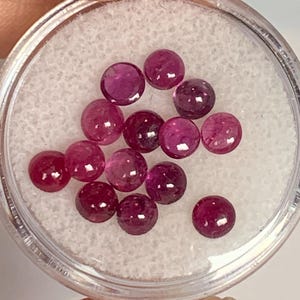 May include: A close-up of a small container filled with 14 round, pink gemstones. The gemstones are smooth and shiny.