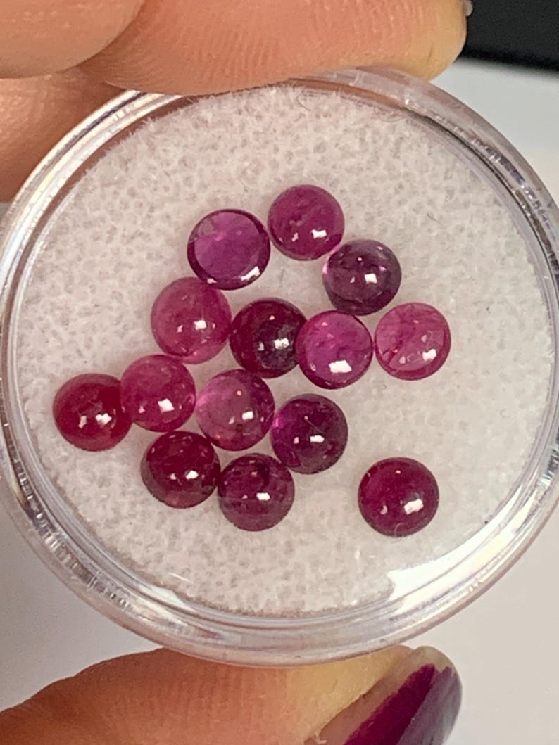 May include: A close-up of a small container filled with 14 round, pink gemstones. The gemstones are smooth and shiny.