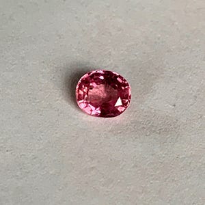 May include: A faceted, oval-shaped, pink gemstone. The gem has a brilliant cut, reflecting light with multiple facets. The color is a vibrant, medium pink, with a slight variation in tone across the surface. The gemstone is set against a neutral background.