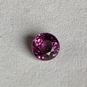 May include: A round, faceted, deep pink gemstone. The gem has a brilliant cut, reflecting light with multiple facets. The stone is set against a neutral background, highlighting its color and clarity. This gemstone is suitable for jewelry.