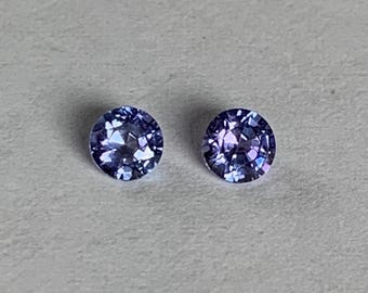 May include: Two round, faceted gemstones with a deep blue-violet color. The gems are set against a light gray background, showcasing their brilliant cut and reflective surfaces. These could be used for jewelry.