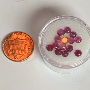 May include: A small plastic container filled with approximately 15 round, pink gemstones. The container is sitting on a white surface next to a US penny.