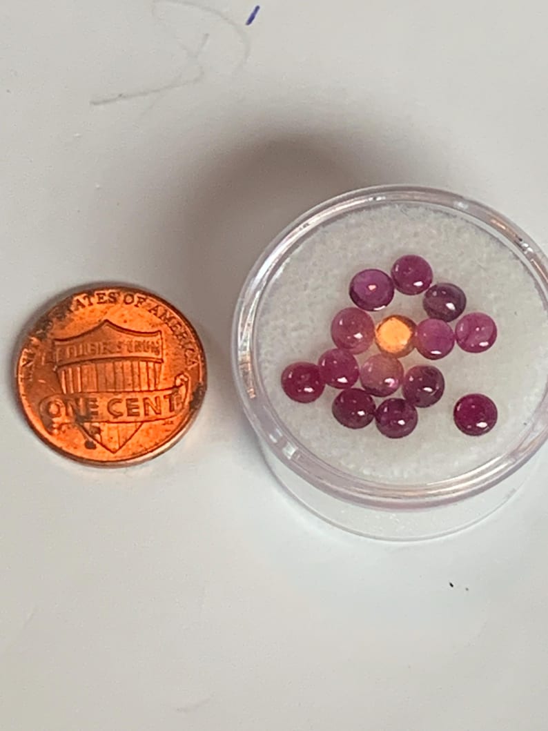 May include: A small plastic container filled with approximately 15 round, pink gemstones. The container is sitting on a white surface next to a US penny.