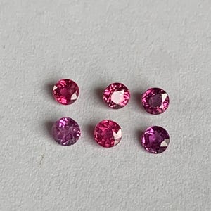 May include: Six small, round gemstones in shades of pink and purple are arranged on a light gray surface. The gems have a faceted cut, reflecting light. The colors range from deep magenta to light pink.