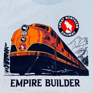 Peut inclure: Un t-shirt bleu clair avec une illustration vintage d'une locomotive, des montagnes et un logo pour le Great Northern Railway. Le texte "Empire Builder" est imprimé sous l'image.