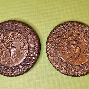 May include: Two antique bronze-colored decorative medallions. Each medallion features a central circular design with a bird and tree motif, surrounded by a floral border. The background is a light green color.