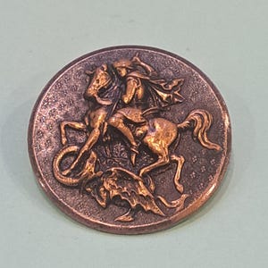 Peut inclure: Une pièce de monnaie ronde, de couleur bronze, avec un relief détaillé d'un chevalier à cheval tuant un dragon. Le chevalier porte une cape et la pièce a un fond texturé.