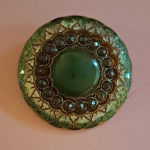 May include: A circular button featuring a smooth, dark green center surrounded by a ring of small, dark brown, faceted beads. The outer edge displays a textured, scalloped pattern in shades of forest green, cream, and metallic gold accents.
