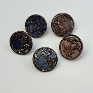May include: Five circular buttons featuring raised metallic floral and botanical designs in bronze tones. The buttons have smooth, colored centers in shades of dark blue, light blue, and muted brown, each framed by a textured, rope-like metallic edge.