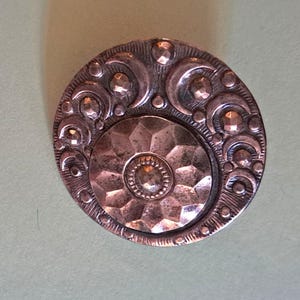 Peut inclure: Un bouton circulaire de couleur cuivre présentant un médaillon central à facettes entouré d'une bordure en relief ornée de formes de croissants de lune, de petits points circulaires et de lignes texturées, créant un motif métallique détaillé.