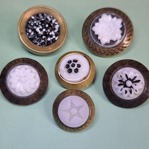 May include: A collection of five circular buttons featuring brass-toned metal rims and decorative centers. The designs include a black and white textured pattern, a white floral relief, a white star, and two white floral motifs with dark accents.