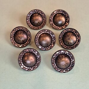 May include: A set of six round, dark bronze-toned buttons featuring a raised, smooth center dome with a subtle etched starburst pattern. Each button is framed by a decorative, textured border with a repeating open-work pattern.