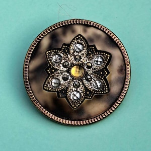 May include: A round button featuring a brown tortoiseshell-patterned base with a metallic gold-toned rim. The center displays a six-petaled floral motif in antique gold metal, accented with small clear crystals and a central yellow stone.