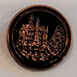 May include: A round, bronze-colored button with a detailed relief of a castle on a rocky landscape. The castle features multiple towers and architectural details. The background is black, and the button has a raised edge.