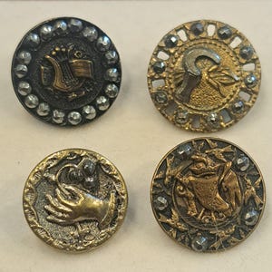 May include: Four circular metal buttons with intricate relief designs. One features a lyre, one a sickle, one a hand holding a flower, and one a bird motif. Each button is accented with small, faceted metallic studs along the outer edge.
