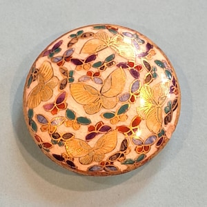 May include: A round, flat, circular object featuring a cream-colored background decorated with multiple gold-toned butterfly outlines. The design includes small, colorful enamel-filled sections in shades of red, purple, green, and blue throughout.