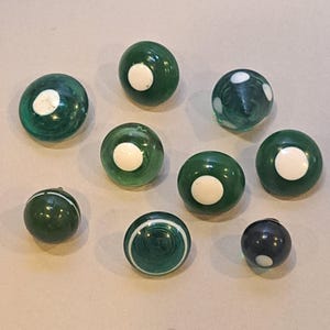 May include: A collection of eight round glass buttons in varying shades of green. Most buttons feature a smooth, translucent green surface with a solid white circular inlay in the center, while others display white circular or linear patterns.
