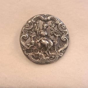 Puede incluir: Un botón metálico circular de tono plateado con una escena en relieve de una figura sentada flanqueada por dos ciervos. El diseño está rodeado por un borde ornamentado con volutas. La palabra MUM está estampada en relieve en la parte inferior.