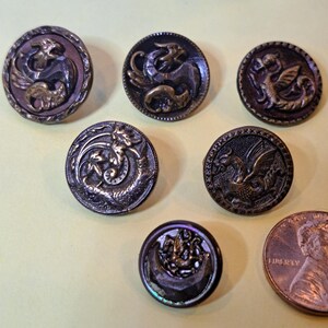 Può includere: Una collezione di sette bottoni antichi in metallo con disegni di draghi. I bottoni sono principalmente in bronzo e marrone scuro, con intricati dettagli in rilievo. Un bottone è iridescente. Una moneta è inclusa per il confronto delle dimensioni.