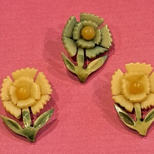 May include: Three flower-shaped pins featuring layered petals and a central bead. One pin is sage green with a green stem and leaves, while the other two are cream-colored with green stems and leaves. Each pin has a distinct, carved-style petal design.
