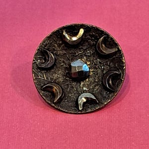 May include: A circular bronze-toned button featuring a textured floral background. The button is decorated with five raised crescent moon shapes arranged in a circle around a central, faceted, dark metallic stone that reflects light.