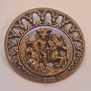 Könnte beinhalten: Ein runder Knopf im antiken Stil mit Bronze-Finish. Der Knopf zeigt ein detailliertes Relief einer Figur auf einem Pferd, umgeben von einem durchbrochenen Rand. Das zentrale Bild zeigt einen Ritter auf einem Pferd, mit einem Drachen unten.