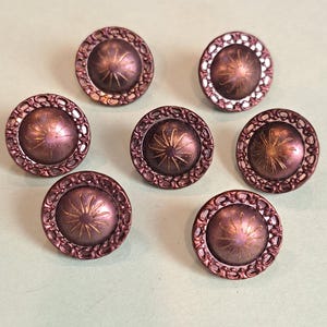 May include: A set of seven round buttons in a metallic plum purple finish. Each button features a raised central dome with a sunburst pattern and a decorative filigree border around the edge. The buttons have a smooth, slightly reflective surface.