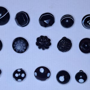 Può includere: Una collezione di bottoni neri di varie forme e dimensioni, tra cui design rotondi, sfaccettati e strutturati. Alcuni bottoni presentano accenti bianchi, strisce o abbellimenti floreali. Una moneta è inclusa per il confronto delle dimensioni.