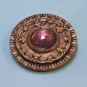 May include: A round copper-toned button featuring a central faceted purple stone. The button has a textured basket-weave pattern in the center, surrounded by an ornate raised border with scrollwork details and a fluted outer edge.