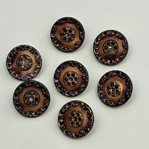 May include: A set of six round buttons in a metallic bronze finish. Each button features a central circular inlay with small embedded crystals, surrounded by a raised, ornate border with intricate scrollwork patterns along the outer edge.