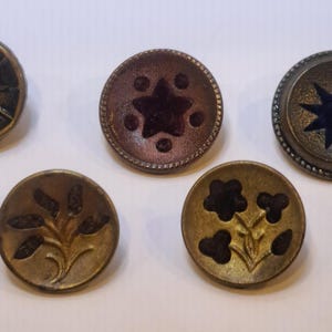 May include: A collection of antique buttons in various designs. The buttons feature star and floral patterns, with colors including bronze, dark blue, and brown. The buttons are round and have a vintage aesthetic.