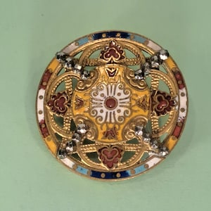 May include: A circular gold-tone brooch featuring intricate openwork metal detailing. The center displays a yellow and white enamel floral motif, surrounded by red, blue, and white enamel accents. Four faceted dark gray beads are set in a cross pattern.