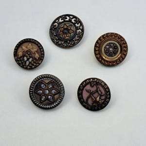 May include: Five circular buttons with metallic bronze and copper tones. Each button features intricate relief patterns, including floral motifs, star shapes, and embedded clear crystals. The designs vary in texture, with some showing smooth centers and others displaying detailed filigree edges.