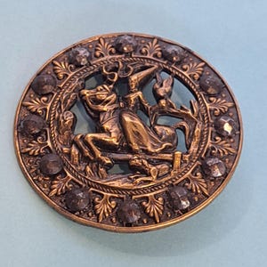 May include: A round, antique brass button with a detailed relief scene. The central design depicts a figure riding a horse, surrounded by foliage and accented with dark, faceted stones around the edge. The button has a vintage aesthetic.