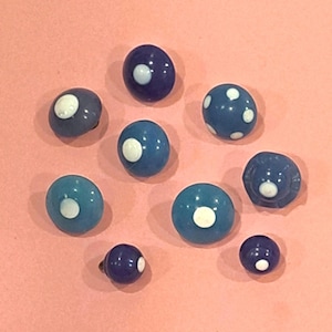 May include: A collection of eight round, dome-shaped buttons in various shades of blue, ranging from light sky blue to deep navy. Each button features a smooth, glossy finish and a single white circular accent centered on the top surface.
