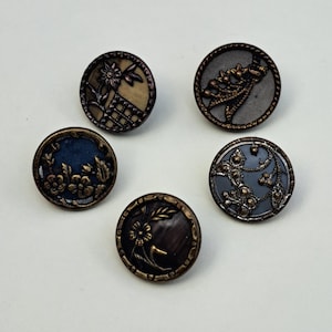 May include: A set of five circular buttons featuring intricate metallic relief designs. Each button displays unique floral or botanical patterns in shades of bronze, dark blue, and grey, with raised edges and detailed textures on the surfaces.