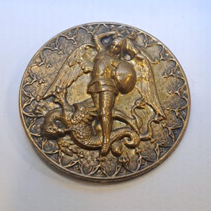 Puede incluir: Medallón redondo de bronce de estilo antiguo. Representa una figura alada, posiblemente un ángel, sobre un dragón. La figura sostiene un escudo y una espada. El borde tiene un diseño floral ornamentado.