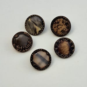 May include: Five circular buttons featuring metallic bronze-toned rims with intricate scrollwork patterns. The centers of the buttons display varied marbled and mottled textures in shades of brown, cream, gray, and black, creating a distinct appearance.